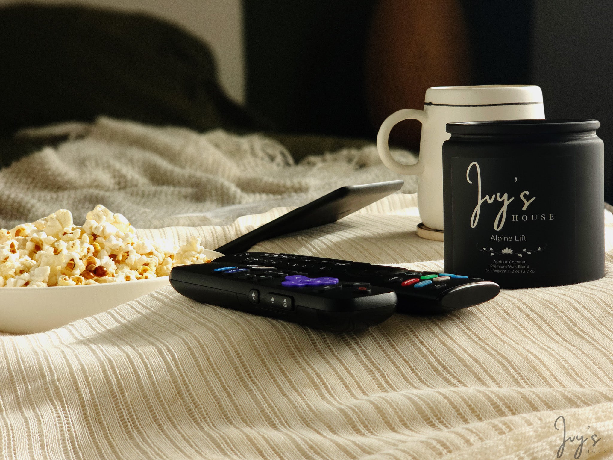 Ivy’s House Alpine Lift candle on a textured table beside a mug, tablet, and remotes in a cozy setting