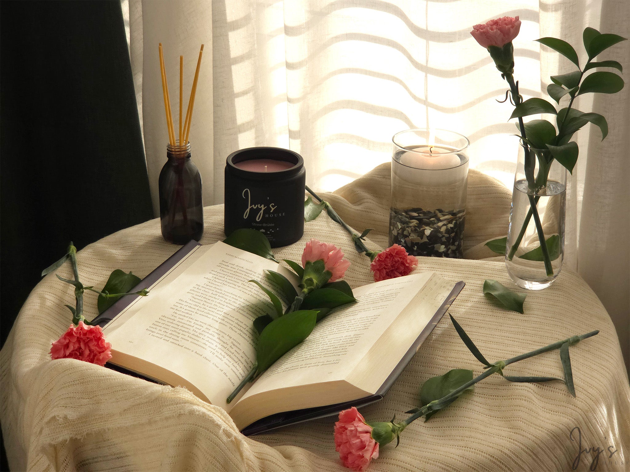 Straw-Brûlée candle by Ivy’s House beside open book, flowers, and lit glass candle in soft light